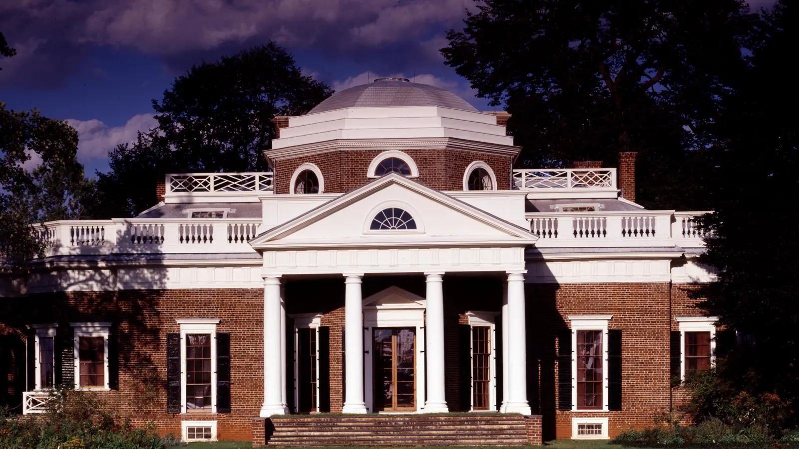 The white-columned west front of Thomas Jefferson's Monticello, with its octagonal dome and red brick walls, under a dusk sky in Charlottesville, Virginia.